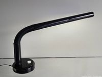 Side profile of the black post modern desk lamp showing its tubular head, flexible ribbed arm, and circular base with switches.