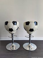 Front view of two matching soccer ball patterned counter stools with chrome bases and footrests, showing the seat shape and adjustment levers.