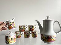 Photo showing the front view of the mini tea set including the teapot, creamer, sugar bowl, and five tea cups. Pieces have colorful flower pattern on white ceramic.
