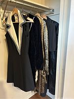 Five vintage women's dresses hanging on wooden hangers on a clothing rack. The front black sleeveless dress features a wide white lapel with black piping. The other dresses vary in colors and textures.