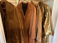 Five various vintage leather and suede coats hanging on wooden hangers in a closet, including a reversible men's suede jacket and a metallic gold coat.