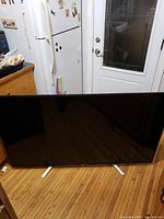 Full front view of the TV showing black screen and silver stands on wooden floor in a kitchen area.