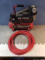 Full view of red Airgo air compressor and coiled red air hose on a white and grey table.
