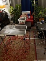 Image showing the camping kitchen table unfolded with side rack extension visible, on red patterned carpet indoors.