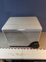 White portable cooler with smooth exterior on floor mat, single black wheel visible at bottom corner