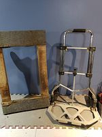 Wooden frame dolly with metal reinforcement and foldable aluminum handcart with plastic platform and rubber handle grip on a blue background.