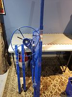 Blue drywall lift standing upright showing the crank wheel and foldable legs on a carpeted floor.