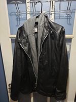 Front view of men's black leather jacket with grey inner hoodie hanging on door hook.