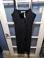 Black sleeveless dress hanging on black plastic hanger against a white framed glass door, showing front view with bodice ribbed and flowy skirt.