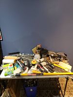 Photo showing assorted painting tools spread on a table against a blue wall, including paint rollers, paint brushes, scrapers, sanding pads, paint trays, and painter's tape.