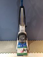 White Hoover upright vacuum cleaner with clear dust container and black power cord, photographed against a blue wall on a foam mat surface.