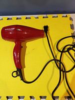 Red Babyliss PRO Volare hair dryer with attached power cord and standard plug, laying on yellow and brown floor mat.