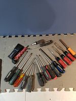 Image shows 13 screwdrivers laid out on gray foam mat including flathead and Phillips heads, with handles of varied colors and materials.