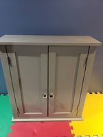 Front view of closed small white wooden cabinet with two doors and metal handles, placed on colorful floor mats in front of blue wall.