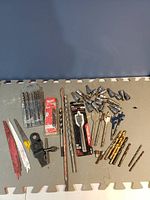 Wide view of lot showing assorted saw blades, multiple sizes of drill bits including spade bits, ratchet driver bits and various ratchet sockets scattered on floor mat.