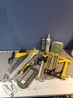 Collection of hand tools including saw, clamps, caulk, mallet, pliers, measuring tape shown on gray mat against blue wall.
