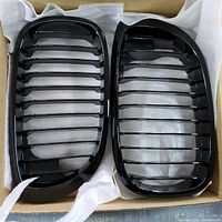 Pair of black BMW kidney grills with horizontal slats, displayed side by side inside a box with protective padding.