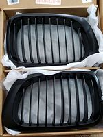 Close-up showing two individual black kidney grills with vertical slats for BMW E46 M3, packed in protective material inside a box.