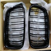 Pair of black glossy BMW front grills with horizontal slats displayed in cardboard box, showing design and finish.