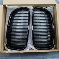 Two black BMW front grilles side by side in a box with protective white paper, showing their kidney shape and horizontal slats.