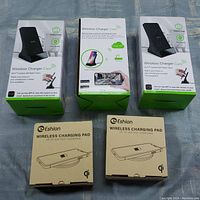 Lot showing three boxed black wireless chargers with integrated WiFi cameras and night vision plus two boxed Eshion wireless charging pads on a surface.