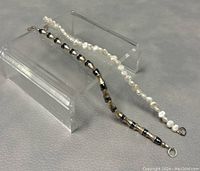 Two bracelets displayed on a clear holder: one white irregular fresh water pearl bracelet and one beaded 925 silver bracelet with alternating barrel and rounded beads.