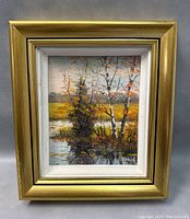 Close view of the framed oil painting showing autumn trees by water, with artist signature visible in bottom right corner and some wear on the gold frame.