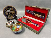 Five-piece vintage Asianware including cloisonné enamel hand mirror and comb set in original green fabric case with red velvet lining, decorative floral and deer enamel plate, colorful floral trinket box, and small Japanese gilt-accented sake cup.