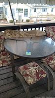 Round aluminum patio table with obscure glass top and umbrella hole. Surrounded by chairs and ottomans with red and beige leaf patterned cushions, beige protective covers seen on deck.