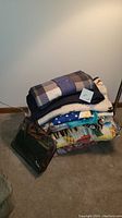 Stack of assorted blankets including a folded blue and gray plaid blanket on top and a brown storage bag on the side