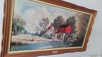 Photo of framed print depicting a red watermill beside a river with a woman in red and white close to the water, surrounded by trees and a white house. Wooden ornamental frame with carved edges visible.
