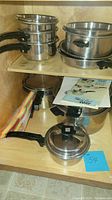 Photo showing multiple FlavorSeal stainless steel cookware pieces stored on wooden shelves, including pots and pans with black detachable handles, lids, a product booklet, and colorful packaging material.