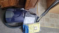 Kenmore Advanced Allergen vacuum cleaner lying on carpet, with vacuum hose and accessory attachments visible. Pack of vacuum bags and printed user manual placed on top.