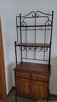 Front view of the kitchen pantry storage hutch showing wood base cabinet with drawers and cupboard, black metal framing above with wooden shelf, wine rack, and hooks.