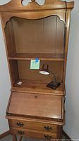 Full view of the secretary desk showing wood finish, desk lamp on the desk's fold-down surface, attached bookcase above with two shelves.