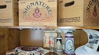 Three wooden crates marked 'Signature Stroh Family brewers since 1775' on a shelf with brewing memorabilia below including glass and decorative beer steins and marketing coasters scattered on the shelf.