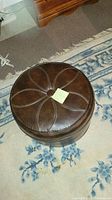 Round brown leather hassock footrest with stitched petal pattern and central button tuft photographed on a carpet with a patterned blue and white rug nearby.