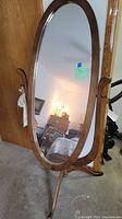 Full-length oval wood frame standing mirror with curved tripod wooden base and side handles for tilt adjustment.