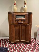 Front view of the antique GE console radio KL-76 in weathered condition with visible scratches and two bowling pins on top, showing the radio dial, knobs, and lower cabinet doors.