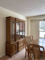 Full view of 2-piece wooden dining hutch with glass upper doors and wooden lower cabinets positioned near window with dining table and chairs.