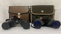 Front view of two pairs of binoculars with their carrying cases behind them on a white surface.