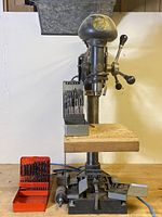 Front view of the Canadian Buffalo drill press with drill bit sets on a wooden workbench, showing the drill press base, wooden table, and accompanying drill bits.