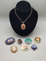 Full view of eight costume jewelry pieces including seven brooches with various designs and one long 1928 pendant necklace on necklace stand.