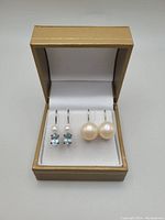 Full view of both pairs of earrings presented in a beige jewelry box with white interior, showing the freshwater pearls and aquamarine stones, respective earring backs visible