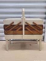 Image shows the closed wooden sewing box with beige legs and wooden brown extending tiers, standing on a flat surface.