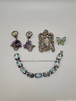 Top-down image showing all items: clip-on earrings, butterfly pin, clip-on picture frame with black and white photo, and Taxco bracelet with blue inlays.