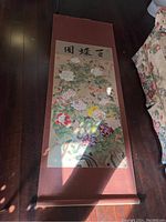 Full view of the Asian paper scroll wall hanging laid flat on the floor beside a floral sofa, showing the full artwork with calligraphy and fabric border.