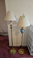 Two standing brass floor lamps next to each other, one with plain brass shaft and white shade, the other with blue decorative section and pleated white shade