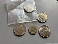 Five British coins lying on a gray surface. Included are two Churchill coins, a sealed coin likely the 1921 florin, the QE2 £5 coin, and Prince Charles and Lady Diana coin.