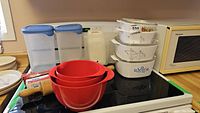 Overview of entire lot on stove showing CorningWare casseroles stacked, Rosti mixing bowls nested, rolling pin, and plastic storage containers.